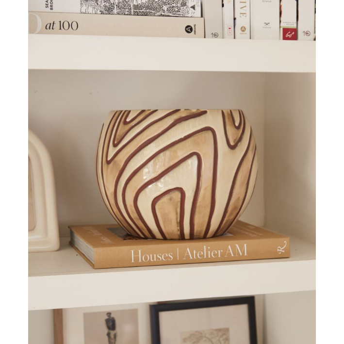 A round, beige ceramic Malina Pot with dark brown wavy lines sits atop a beige hardcover book titled "Houses | Atelier AM" on a white shelf. The pot's contemporary ceramic design adds functional and stylish flair to the neatly arranged books and decor items on the surrounding white shelves.