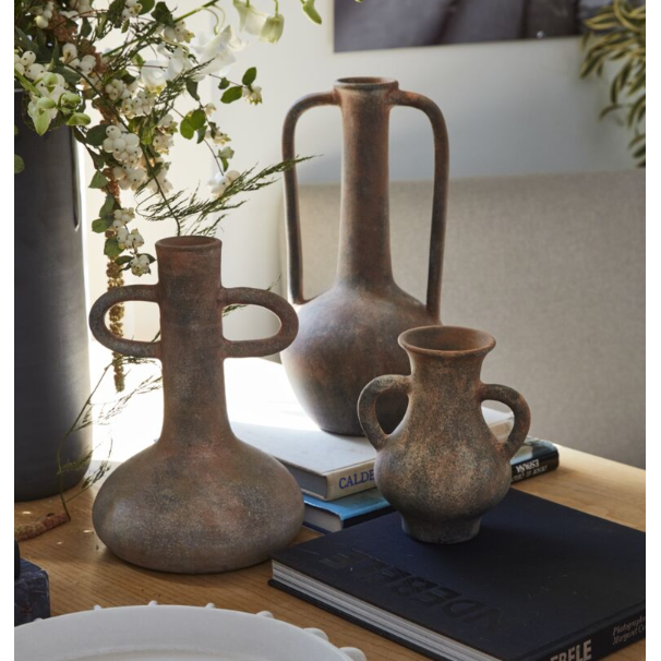 The Tupita Vase, featuring a rustic, sculptural design with a unique handle and weathered terracotta texture, sits on a wooden table near books and greenery. Sunlight highlights its details; in the background, a gray couch and framed photo are visible.