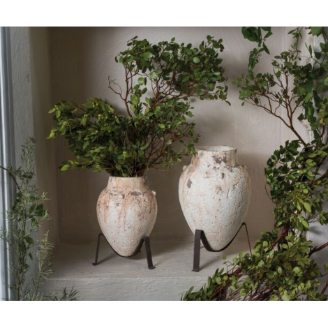 On a ledge, two Tuscan Plant Stands featuring rustic, handmade clay pots create an artisanal charm. The pot on the left is filled with lush green foliage, while the one on the right remains empty. Set against a light-colored wall and framed by partial greenery, they evoke a serene and natural Tuscan ambiance.