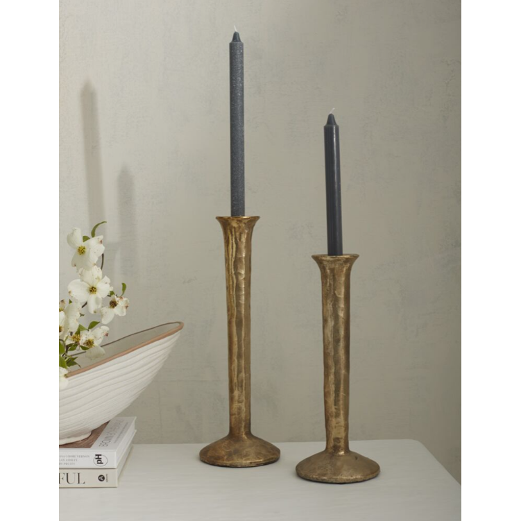 A white tabletop displays two Celestial Gold Candlesticks, each holding a tall, unlit black candle. The candlesticks feature a stunning mottled gold finish and are of different heights, with one being taller than the other. Beside them sits a decorative white bowl with brown trim, filled with white flowers. The backdrop is a plain, light-colored wall.
