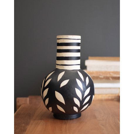 Ideal for enhancing the aesthetic of a home or office, the Black and White Paper Mache Bottle Vase is crafted from black ceramic, adorned with graceful white leaf designs. The neck features an attractive pattern of black and white horizontal stripes, reminiscent of classic paper mache art. Placed on a wooden table, it complements a stack of books and stands out elegantly against a dark gray wall.