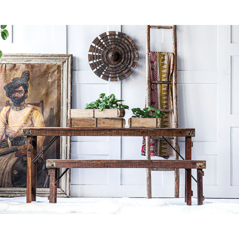 A Collapsible Wedding Bench with folding legs sits against a white paneled wall. Above, a circular decor piece hangs near a ladder with patterned textiles, evoking Indian weddings' charm. A large vintage painting leans against the wall, surrounded by crates and green plants.