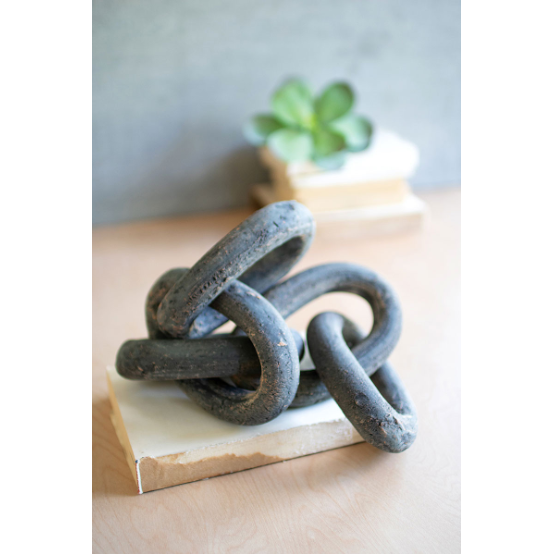 A distinctive Lost Link Clay Chain, with its large, interwoven black rings resembling a unique chain design, is elegantly displayed on a white rectangular base. In the background, a small green succulent plant sits atop two stacked books on a wooden surface, completing an elegant tablescape.