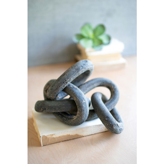 A distinctive Lost Link Clay Chain, with its large, interwoven black rings resembling a unique chain design, is elegantly displayed on a white rectangular base. In the background, a small green succulent plant sits atop two stacked books on a wooden surface, completing an elegant tablescape.