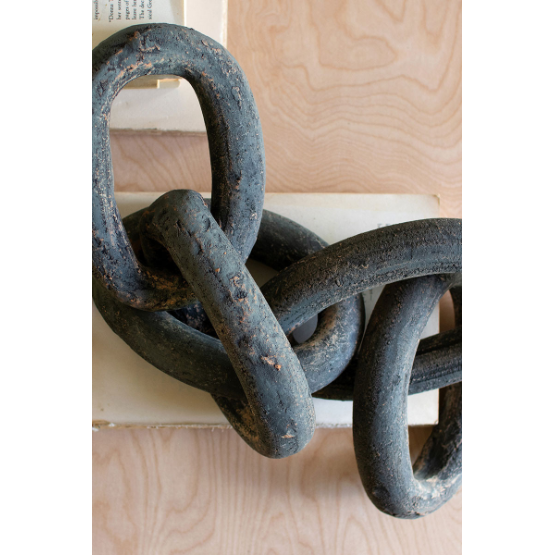 Close-up of the Lost Link Clay Chain with its distinctive, heavy, weathered black metal links arranged on a light-colored wooden table. The large links exhibit signs of rust and corrosion. A portion of a white object is partially visible beneath the chain, set against the backdrop of wood grain texture.