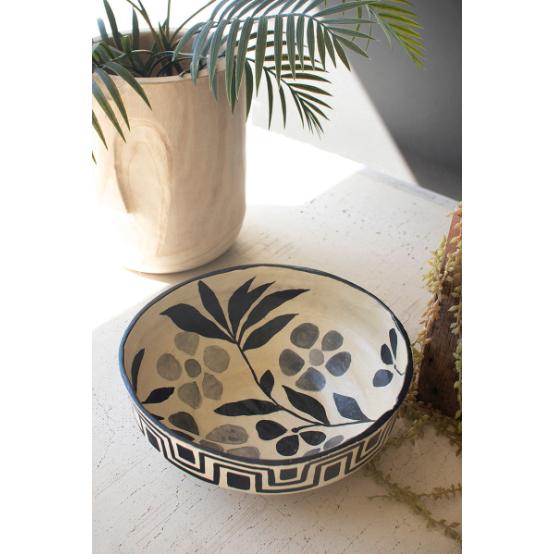 A Black and White Paper Mache Bowl with a black and white floral pattern adorns a light-colored table, exuding sophistication. This elegant bowl showcases black leaves and berries on a white background, complemented by an intricate geometric border. In the backdrop, there's a light wooden plant pot housing a fern, with another plant slightly visible.