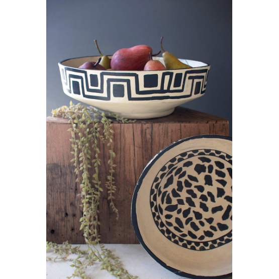 A Black and White Paper Mache Bowl, adorned with a geometric pattern and filled with vibrant red and green pears, rests on a rustic wooden block. Hanging plant tendrils elegantly drape over the block, enhancing its charm. Nearby, another shallow ceramic bowl featuring a black and white speckled design is displayed, leaning gracefully against the wooden structure.