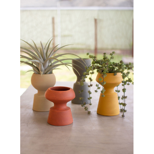 Four differently colored Nell Clay Vases sit on a light-colored surface, with houseplants in three of them. From left to right, the vases are terracotta with an air plant, beige and empty, light gray with a succulent, and mustard yellow with a trailing succulent. They could easily double as stylish clutter catchers. A blurred background is visible.