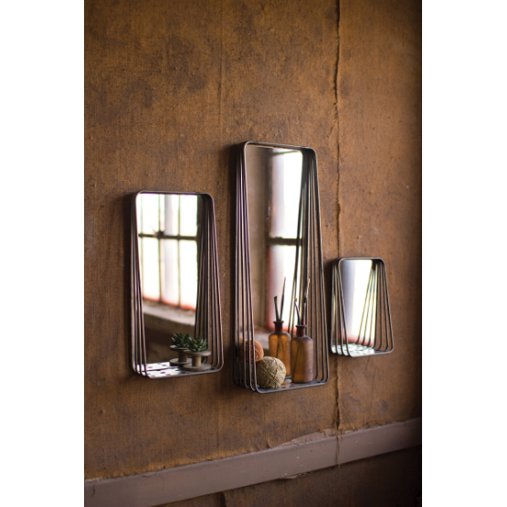 Three wall-mounted Metal Framed Mirrors with wire frame edges reflect a dimly lit room. Each mirror varies in size, with the largest holding small decorative items like vases and a succulent on its convenient built-in shelf. The background features a rustic, textured brown wall, adding a vintage feel to the modern home decor scene.