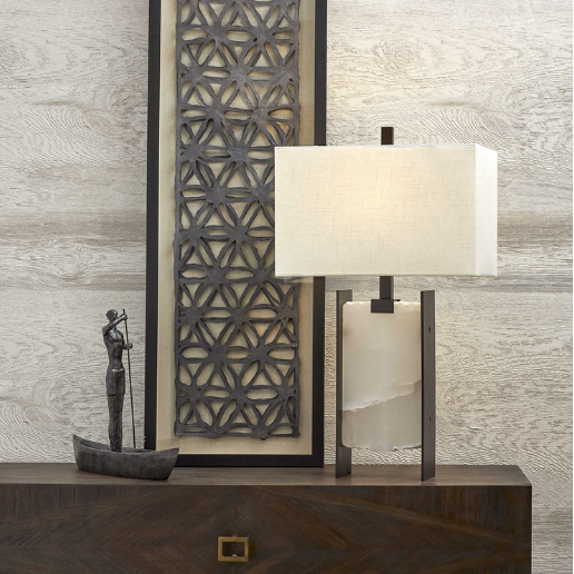 A person standing in a boat makes for a decorative sculpture placed on a dark wooden cabinet that features a square gold handle on the front drawer. Close by, there's a rectangular table lamp with a marble-like base and fabric shade emitting a warm glow. The intricate Evander Wall Art piece behind them enhances the scene.