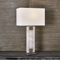The Calista Table Lamp exudes modern elegance with its rectangular white shade, perched on a dark wooden surface against a textured brown wall. The base is crafted from alabaster-like translucent material, highlighted by two dark metallic accents that seamlessly connect it to the shade.