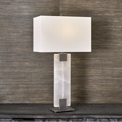 The Calista Table Lamp exudes modern elegance with its rectangular white shade, perched on a dark wooden surface against a textured brown wall. The base is crafted from alabaster-like translucent material, highlighted by two dark metallic accents that seamlessly connect it to the shade.