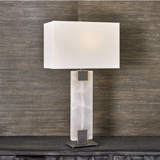 The Calista Table Lamp exudes modern elegance with its rectangular white shade, perched on a dark wooden surface against a textured brown wall. The base is crafted from alabaster-like translucent material, highlighted by two dark metallic accents that seamlessly connect it to the shade.