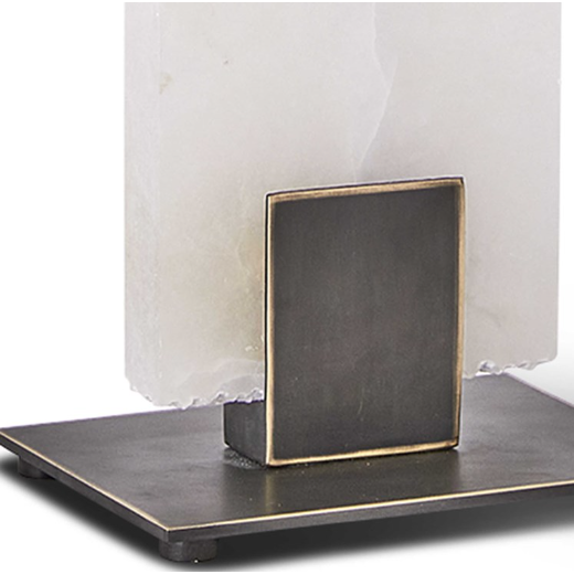 A close-up of the Calista Table Lamp base displays a sleek, dark metallic rectangle adorned with a square, brass accent near the bottom. The lamp's body is crafted from translucent alabaster featuring an irregular edge at its base. This sophisticated piece rests gracefully on a flat, dark metallic plate.