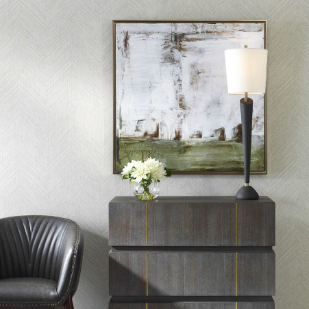 A modern interior boasts a dark wooden chest of drawers adorned with gold accents, topped with a slender black lamp and a small vase containing white flowers. Above it, the Seraphina Wall Art—an abstract framed painting in hues of white, green, and brown—is displayed on the wall. To the left, a black leather chair is partially visible.