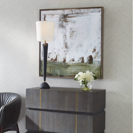 A modern interior features a dark wooden chest with two drawers, accented by thin gold lines. Atop the chest sits a vase filled with white flowers and a sleek black table lamp that has a white shade. The wall behind, adorned in light patterns, showcases the elegant Seraphina Wall Art, enhancing the minimalist abstract painting.
