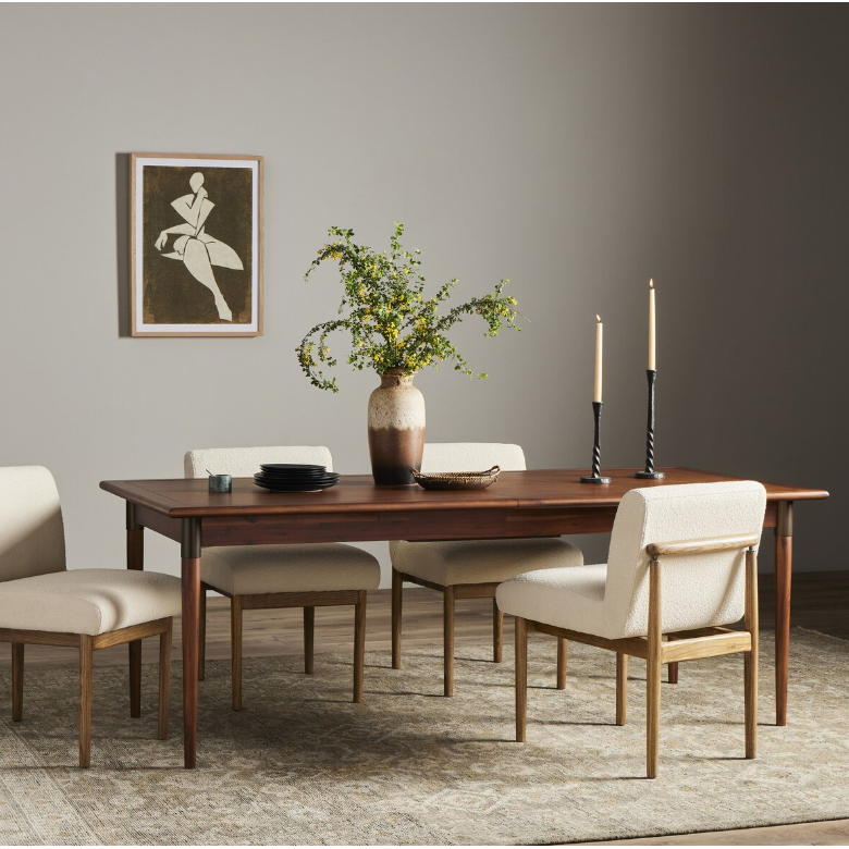 The Teresa Extension Dining Table, crafted from acacia wood, boasts a rectangular surface styled with a large ceramic vase holding green and yellow flowers. It is complemented by two black and white striped candles and a few black dinnerware pieces. The table is surrounded by four cushioned chairs with wooden legs. A framed abstract artwork hangs on the wall in the background.