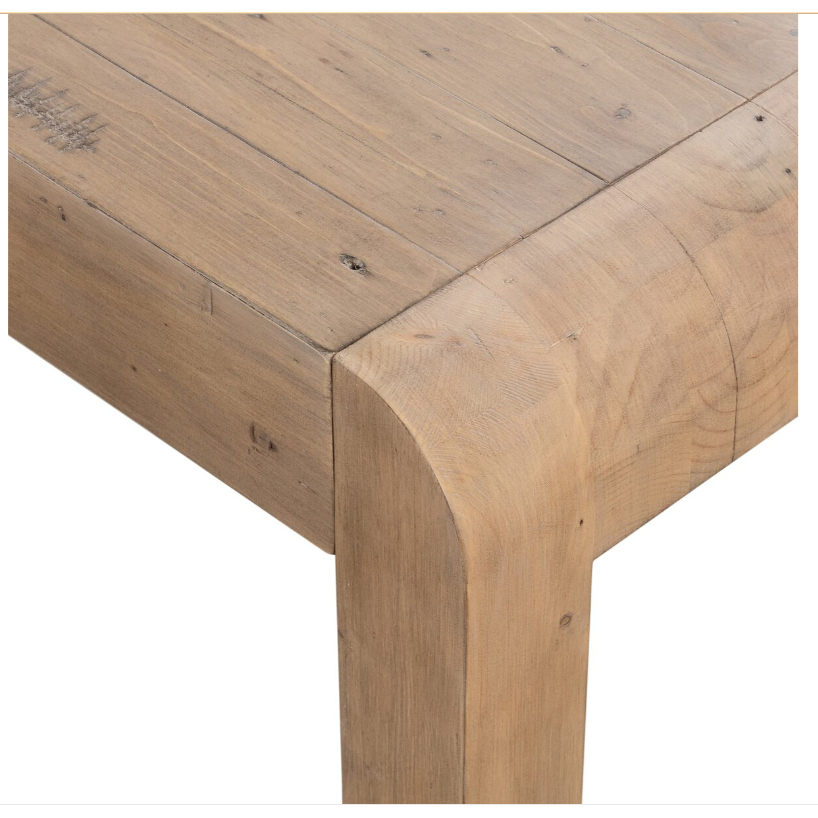 A close-up of the Lotta Extension Dining Table corner highlights its natural, light brown finish with noticeable grain patterns. The table features thick, sturdy legs with rounded edges, seamlessly connected to the tabletop. Small knots and variations in the reclaimed wood further enhance its rustic charm against a white background.