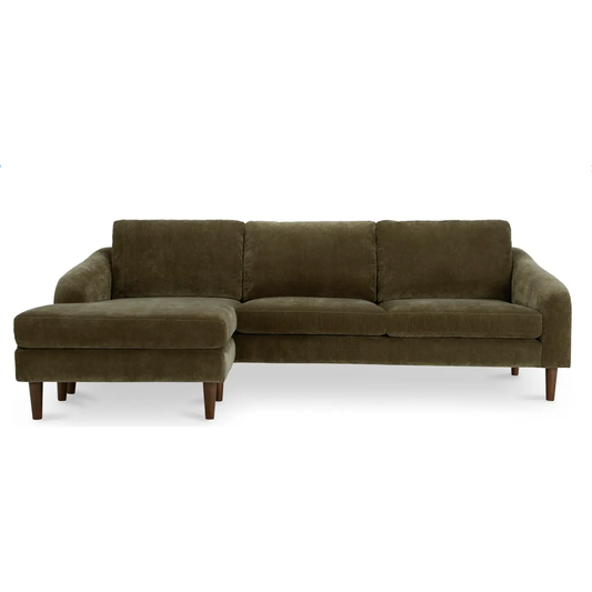 The Ronald Sectional Cedar Green is a dark green, velvet stain-resistant sofa with wooden legs. The modern design features a chaise lounge on the left side, and its plush backrest and seat cushions promise comfort. Positioned against a plain white background, this eco-friendly sofa adds elegance to any space.