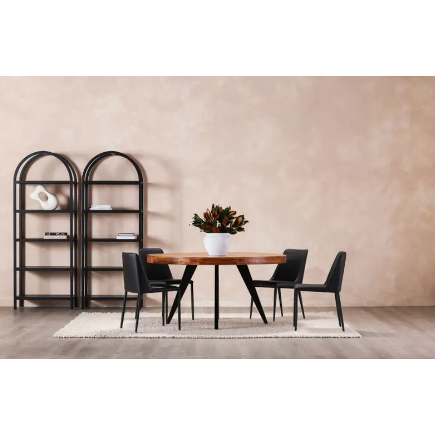 In a dining room, a round wooden table is accompanied by five Nadia Dining Chairs, each upholstered in vegan leather. The setup is completed by a light gray rug underfoot. A white vase filled with deep red and green foliage sits atop the table as a centerpiece. Along the beige walls, two black arched bookshelves display books and decorative pieces, contributing to the contemporary decor.