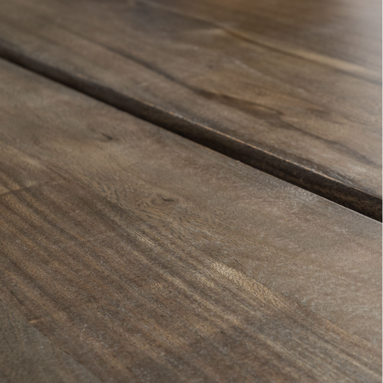 Close-up view of the Vesper 84" Dining Table made from solid Indian mango wood, showcasing its texture and grain. The wooden planks are aligned side by side with visible seams between them, featuring a natural, slightly weathered finish in shades of brown that highlight the organic patterns and character.