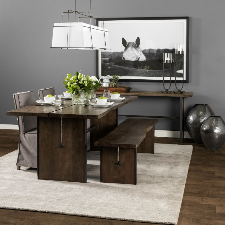 Modern dining room featuring the Vesper 84" Dining Table, crafted from Indian mango wood, adorned with stylish tableware and lush greenery. The seating ensemble includes a matching bench and gray upholstered chairs. A large framed horse photo accentuates the gray wall. A pendant light is suspended above the table. Additionally, two large vases and a candle holder are elegantly placed on a side table.