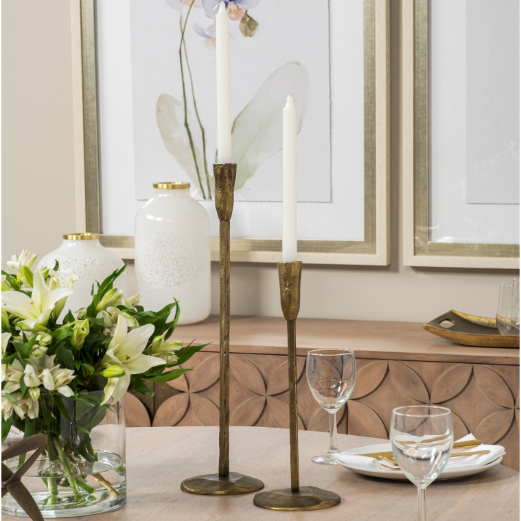 A dining table exuding rustic elegance showcases two Levi Gold Iron Candlesticks gracefully holding white candles. A vase with white flowers and green leaves adorns the left side. The table is set with glassware, a white plate, and cutlery. In the background, floral pictures and a decorative vase complete the scene.