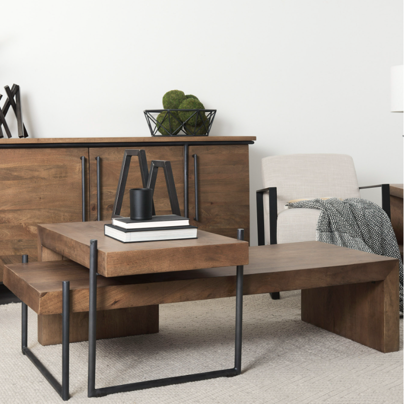 A modern living room features the Thomas Nesting Coffee Tables, expertly crafted from solid Mango wood and supported by sleek U-shaped iron legs. Stacking books and a black candle adorn the tables. In the background, a wooden sideboard with black handles is topped with a decorative bowl filled with green spheres. To the right, a beige armchair draped with a gray throw completes the cozy scene.