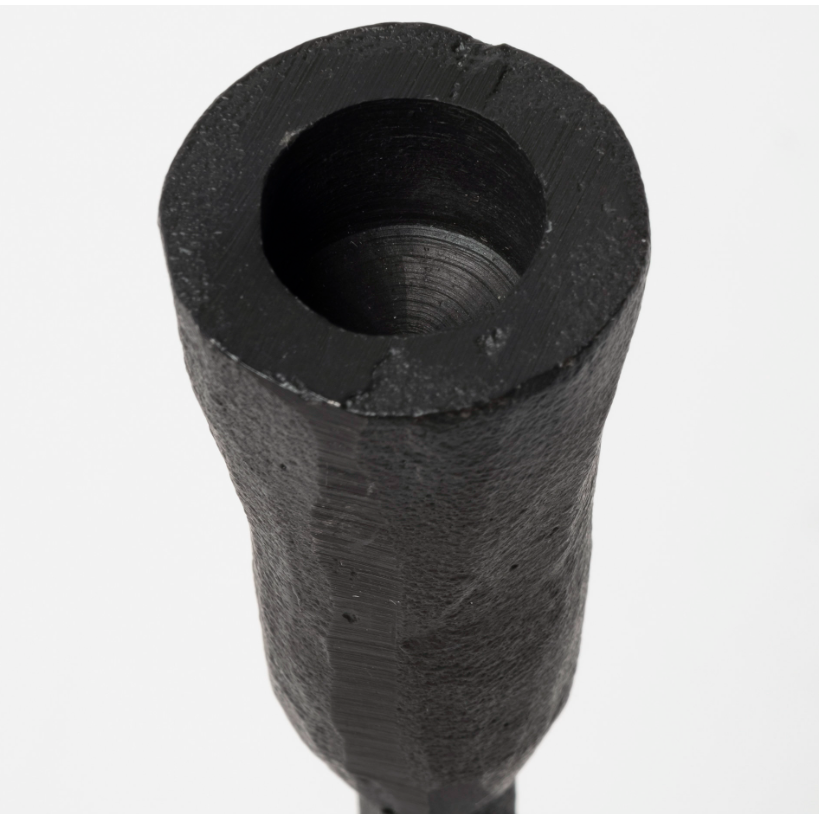 Close-up image of the Levi Iron Candlestick, showcasing its stylish design with a black, textured cylindrical body featuring a hollow, circular opening at the top. The candlestick's surface appears uneven and rugged, exuding rustic elegance with small chips or imperfections near the rim of the opening. The background is a plain, light color.