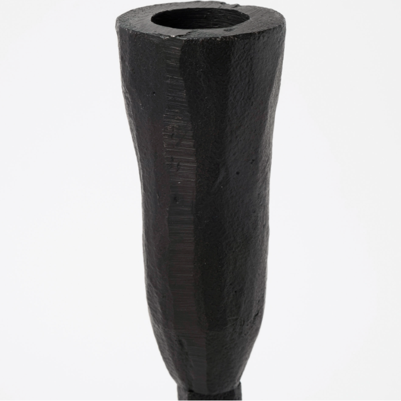 The image showcases a close-up of the Levi Iron Candlestick, a dark, slightly rough-textured sculptural object. It has a cylindrical shape that tapers upwards and ends with a hollow, circular top. The candlestick exudes rustic elegance with carved, uneven finishes and resembles a stylish candle holder against a plain white background.
