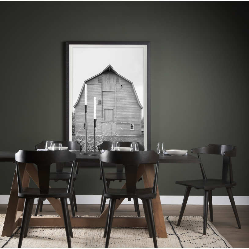 A modern dining area features a dark wooden table with mismatched black chairs set for four. A large, black-and-white framed photo of a barn dominates the wall behind the table. The Levi Iron Candlestick adds rustic elegance to the setting, while dark walls contrast with a light-patterned rug on the hardwood floor. Minimalist decor creates a sleek, contemporary atmosphere.