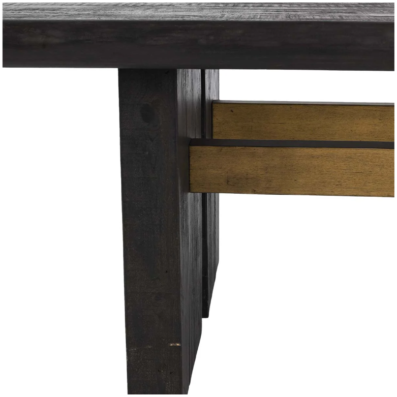 A close-up view of the Echo Reclaimed 96" Dining Table reveals a thick, dark wood leg paired with a horizontal gold-colored metal support beam at the corner. The table's edge showcases a rough texture that beautifully highlights the natural grain of the reclaimed pine wood. This simple, sturdy design accentuates the table's rustic and industrial aesthetic.