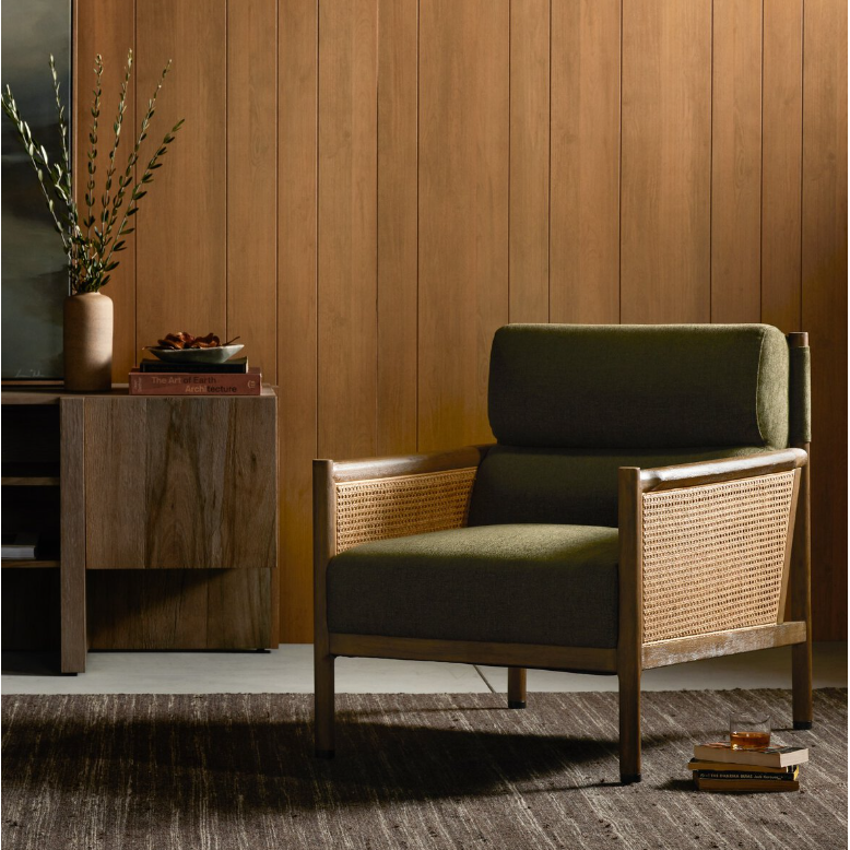 A chic living room features the Julius Accent Chair, with a green cushion and rattan frame, adding French flair against a wooden-paneled wall. It rests on a dark rug. On the left, a wooden cabinet holds greenery in a vase and stacked books, with more books scattered beside the chair.