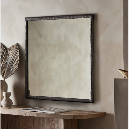 A Hammered Nickel Wall Mirror hangs on a beige wall. Below it, a wooden console table with a natural finish holds a closed book, two dried palm leaves in a textured vase, and another smaller decorative piece beside it. The home decor exudes a minimalist, earthy aesthetic.