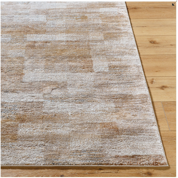 A close-up of a corner of the Sahara Machine Woven Rug reveals its rectangular, textured design on a wooden floor. Featuring a subtle patchwork in earthy tones of beige and brown, this rug combines durability with a warm, cozy appearance. Its color palette is beautifully complemented by the light-finished wooden floor.