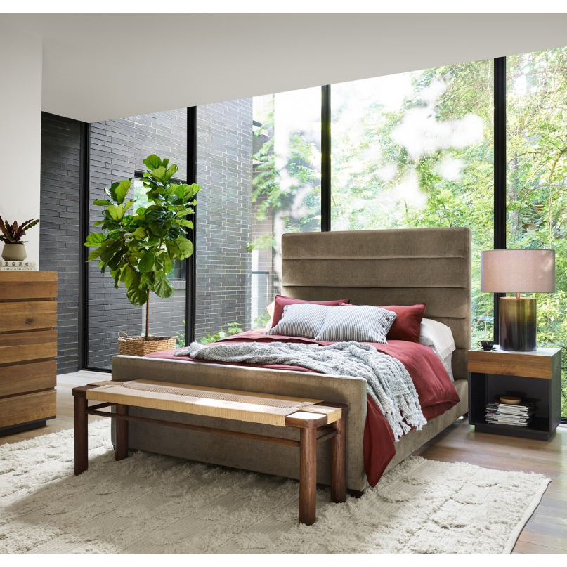 A contemporary bedroom with a large window offers views of lush greenery. The Seraphine Bed features red and gray bedding, boasting a plush upholstered headboard, with a wooden bench positioned at its foot. Beside the bed, there's a wooden nightstand accompanied by a lamp. A potted plant and wooden dresser enhance the decor, harmonizing beautifully with the light-toned rug and flooring.