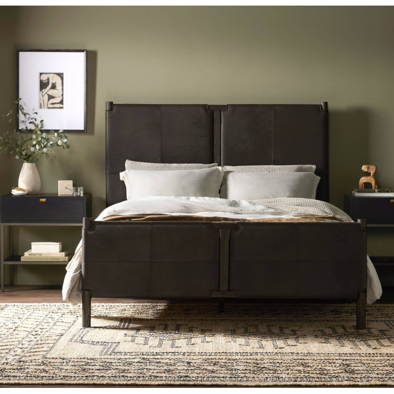A modern bedroom highlights safari-inspired elegance featuring the dark brown upholstered Valencia Haven Bed, complemented by light beige bedding and gray pillows. On either side, black nightstands with lamps enhance the setup. The olive green wall is adorned with an abstract art piece, while a patterned rug ties everything together.