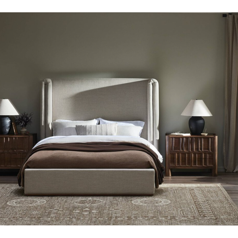 A bedroom beautifully presents the Celeste Bed, featuring its distinctive shelter-style headboard, elegantly dressed in white linens and a cozy brown blanket. The bed is accompanied by wooden nightstands complete with lamps and decorative accents, all situated on a natural wood plinth base. A patterned rug graces the floor, while beige walls and curtains contribute to the warm and inviting ambiance.