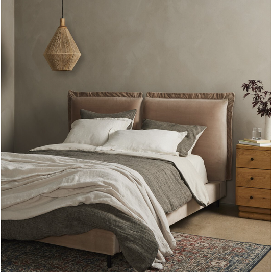 Transform your bedroom into a luxurious retreat with the Ashford Estate Bed, featuring a cozy headboard in beige and gray tones. This elegantly made bed, draped in stylish gray and white bedding, stands out against a textured wall. A woven pendant light illuminates the space from above, while to the right, a wooden nightstand adorned with a plant complements the patterned rug beneath.