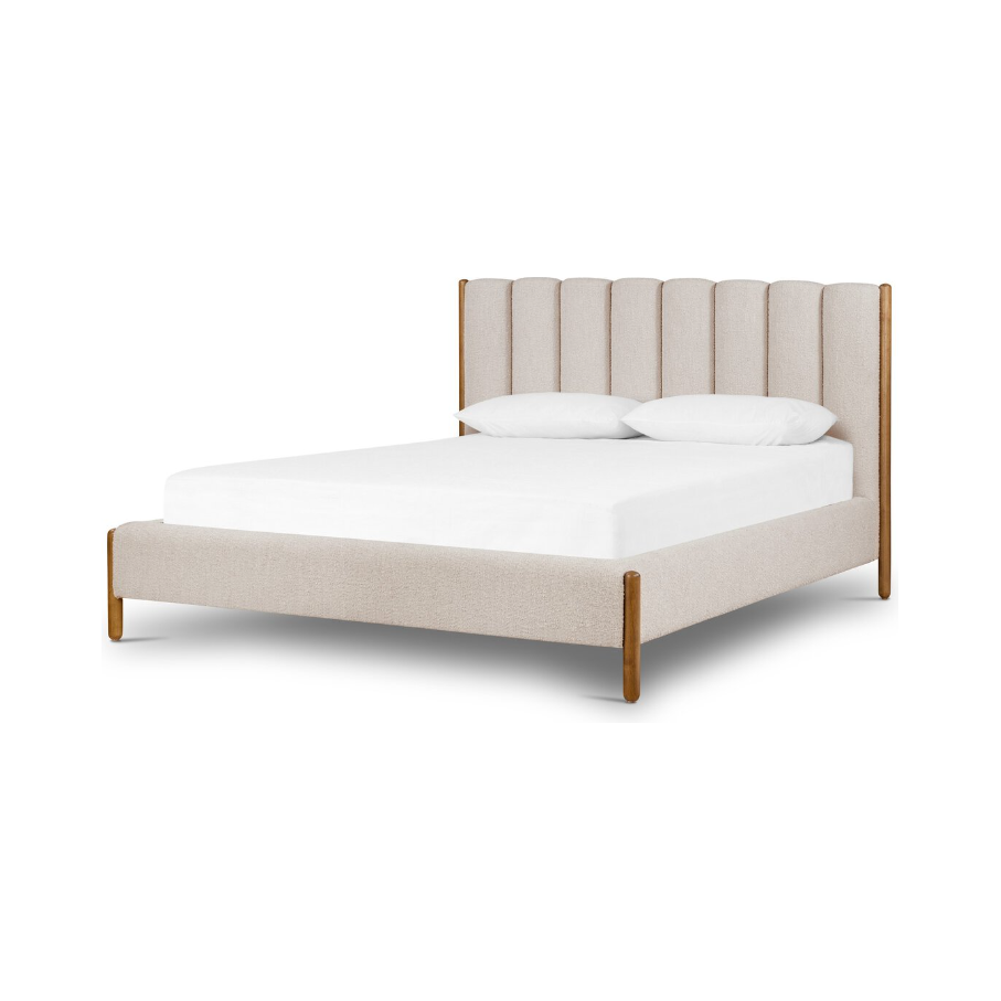 The Savannah Bed is a chic and contemporary upholstered bed in beige, characterized by its tall headboard with vertical panels and simple wooden legs. It comes complete with a neatly arranged white mattress and two white pillows, highlighting the soft textiles alongside its warm wood finish for an elegant touch in modern bedroom furniture.