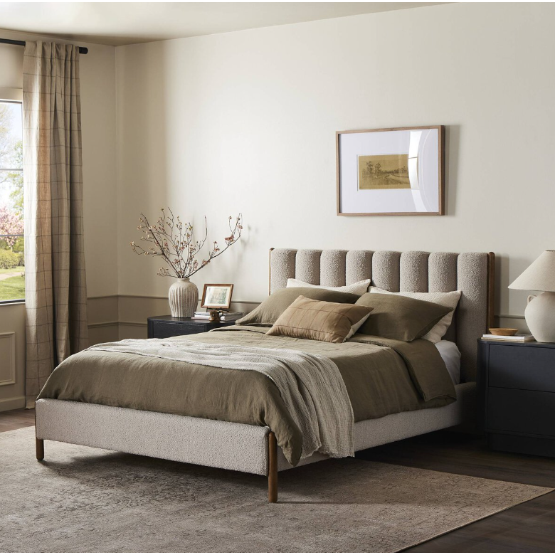 A cozy bedroom highlights the Savannah Bed as the focal point, featuring beige upholstery combined with an earth-toned bedspread and pillows. Flanking the bed are two black nightstands that emphasize modern bedroom furniture styling, each adorned with decor. Above the headboard hangs a framed picture, and sheer curtains permit natural light to illuminate a delicate branch arrangement.