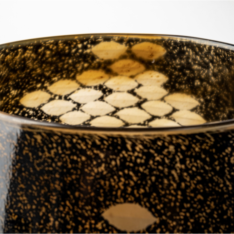 A close-up image of the Marina Black and Gold Glass Vase showcases its cylindrical shape, with a speckled pattern of dark spots. Inside, honeycomb-like formations are visible through the curved surface. Warm lighting casts a golden hue on its elegant design, enhancing its artistic appearance.