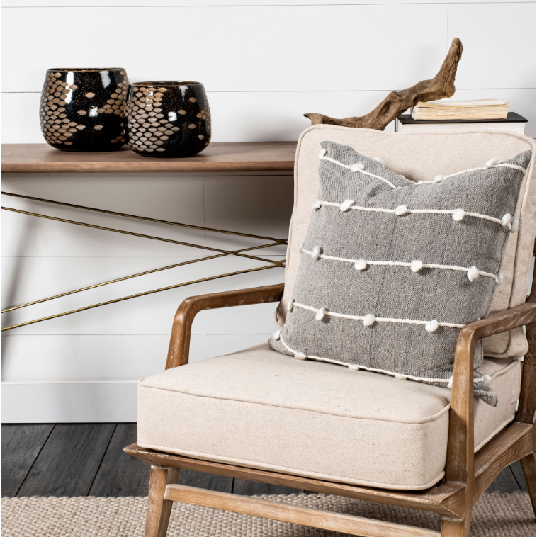 A cozy living space features a wooden armchair with beige cushions and a gray pillow adorned with white pom-poms. Behind the chair, an elegantly designed console table showcases the Marina Black and Gold Glass Vase, decorative vases, and books. The room has white shiplap walls and a dark wood floor.