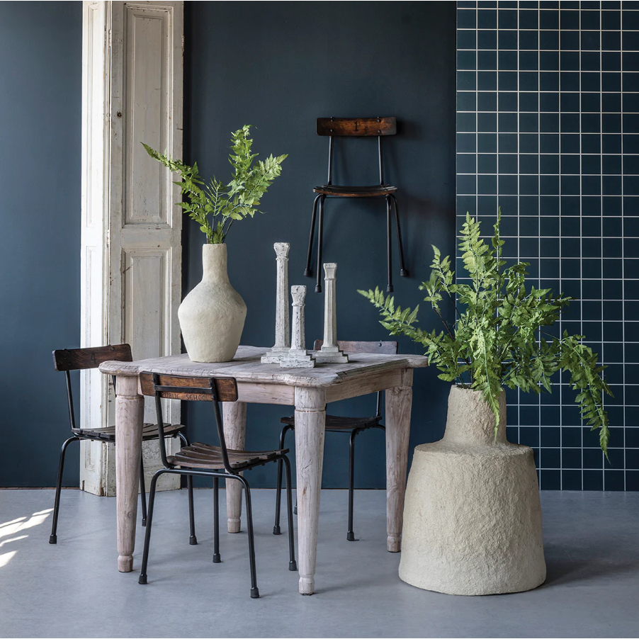 A rustic wooden table with three mismatched chairs holds two tall candlesticks and a Paper Mache Vase with greenery, set near a large planter of ferns. Dark blue walls, white door, and blue tile grid add to this neutral home decor scene.