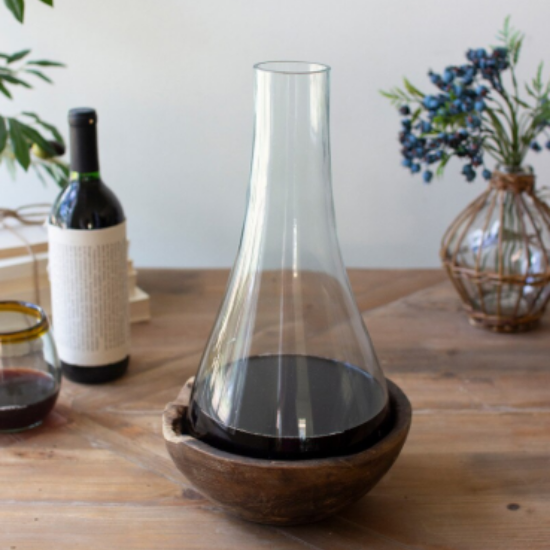 The Teakwood & Blown Glass Bottle with red wine sits in a round teakwood base on a wooden table, alongside a wrapped wine bottle, a glass of red wine, stacked books, and a vase with greenery and purple berries.