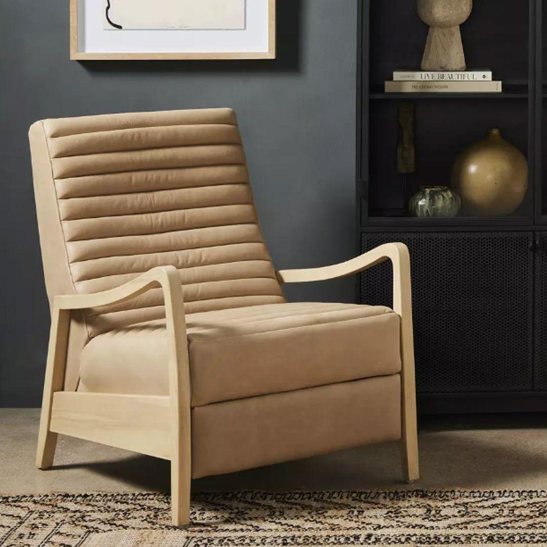 The Terra Leather Recliner in Nude, featuring horizontal tufting and light wood arms and legs, is placed on a patterned rug with a dark wall, framed abstract art, and a black shelving unit with decor behind it.