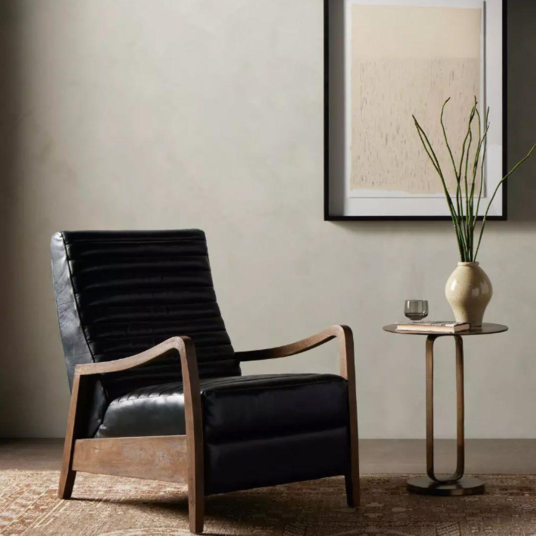 A modern interior showcases the Terra Leather Recliner in black with wooden arms and legs on a textured rug. Beside it, a round side table displays a beige vase with green stems and a glass. Abstract art decorates the neutral wall behind.