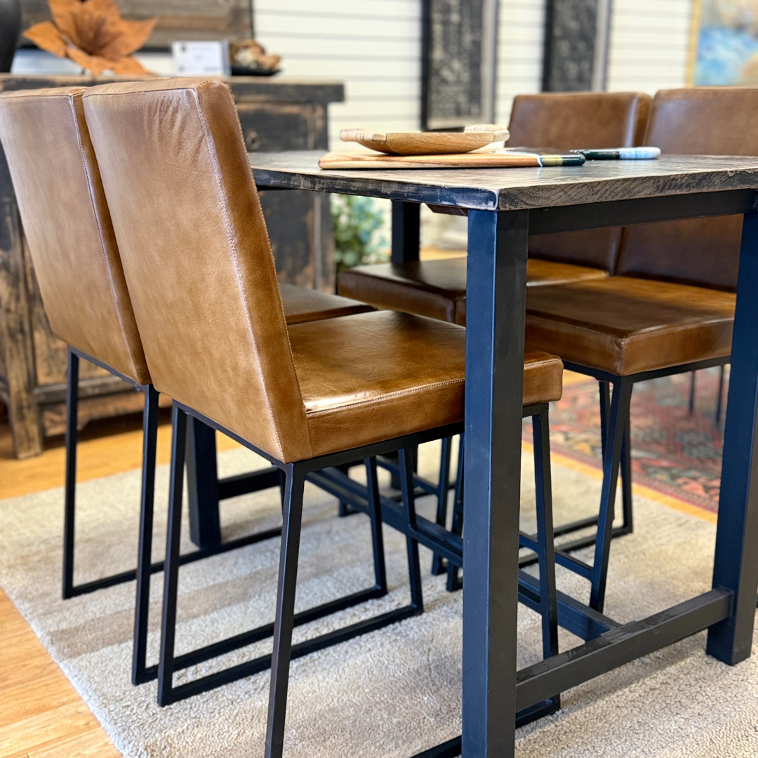 Four Theodore Leather Counter Stools with iron bases surround a dark wooden dining table with a black metal frame. Plates and utensils sit on top, all arranged on a light beige rug in a rustic room with wooden flooring.