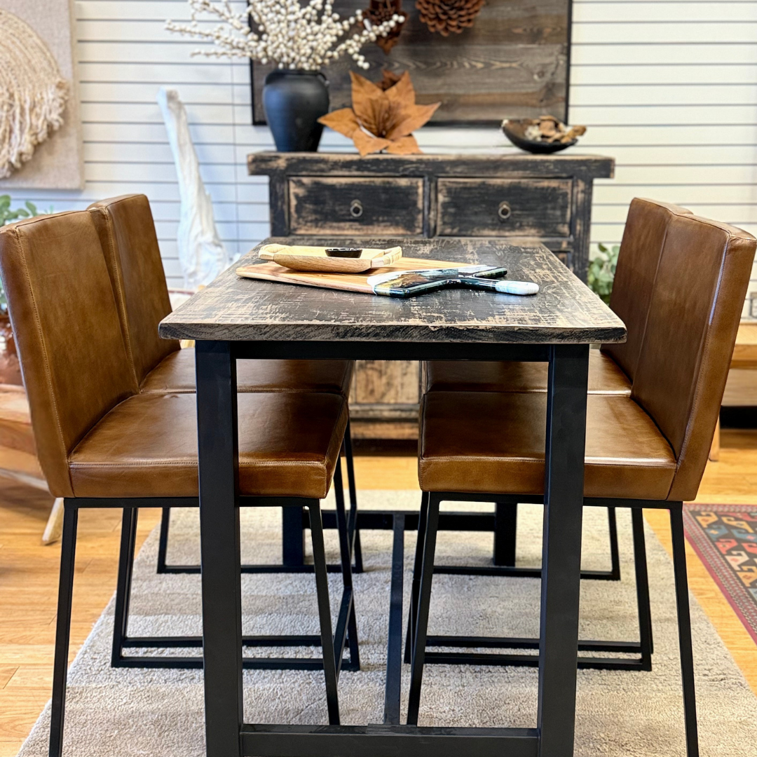A rustic dining setup features a wooden table, four Theodore Leather Counter Stools with cushioned seats, and a textured beige rug. A distressed wood sideboard with decor stands against a white paneled wall in the background.
