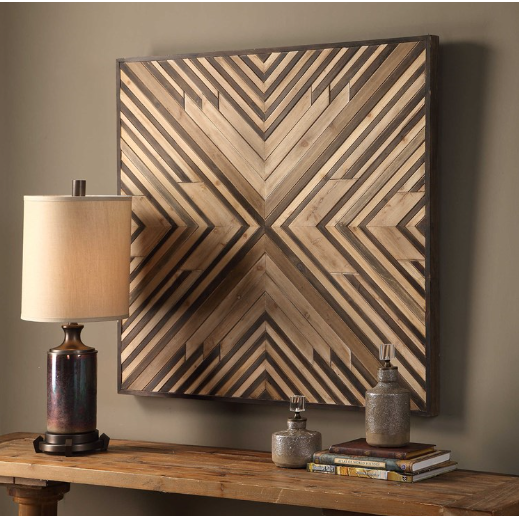 A Thorin Wood Wall Decor with a geometric pattern hangs above a wooden console table, embodying the rustic modern style. The handmade wall art features converging diagonal stripes creating a symmetrical design. On the table, there is a beige lampshade on a dark lamp base, two textured bottles, and a small stack of books.




